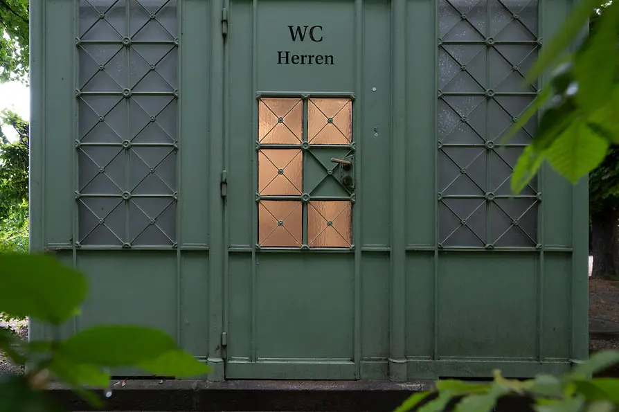 A green historic men’s restroom kiosk with decorative metalwork and frosted windows, partially hidden behind lush leafy branches in a park setting.
