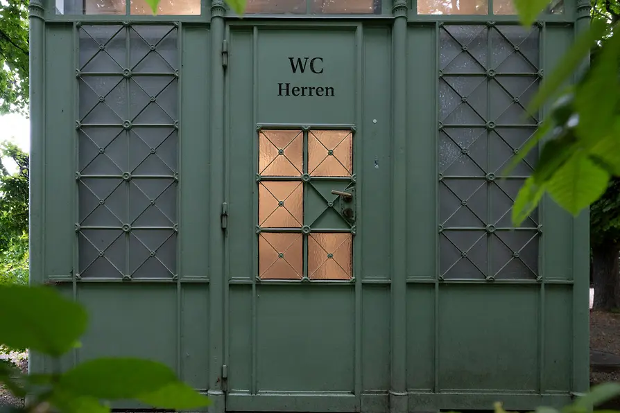 A green historic men’s restroom kiosk with decorative metalwork and frosted windows, partially hidden behind lush leafy branches in a park setting.