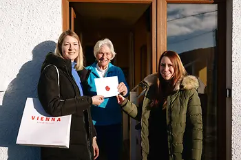 “Three women stand smiling at the entrance of a house; the older woman in the middle holds an envelope sealed with red wax, while one of the women on the left carries a ‘Vienna Now Forever’ gift bag.”