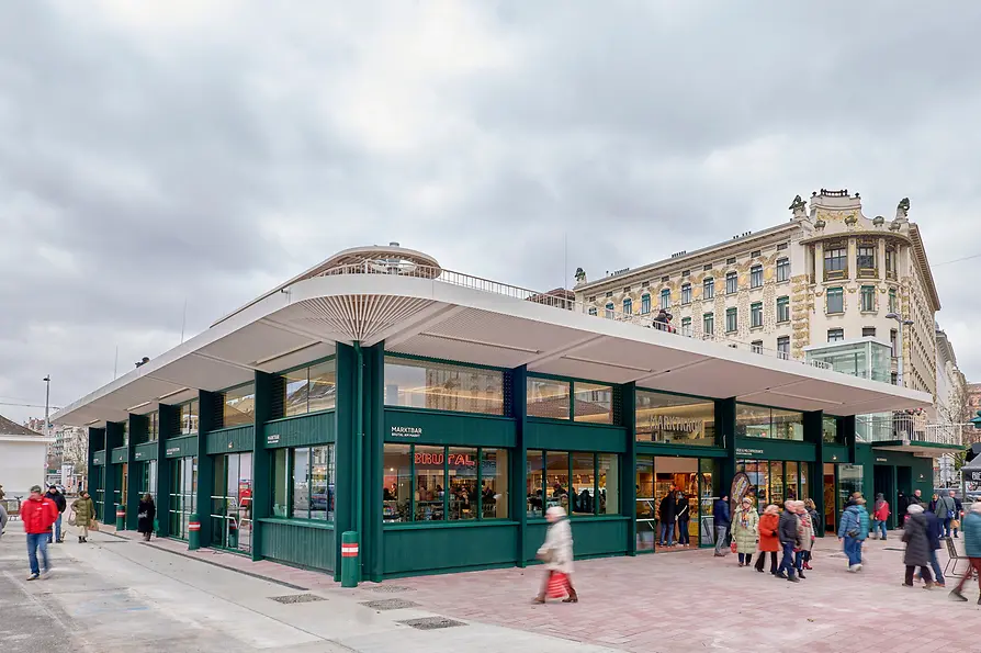 A modern market hall with a wide white roof and green structural elements stands at the Naschmarkt in Vienna. Large windows reveal shops inside, and several people walk by in front of the building. Historic apartment buildings rise behind it under a cloudy sky.