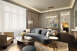 A luxurious hotel suite with a spacious living area featuring a curved blue sofa, light armchair, round coffee table, and soft beige carpet. Large windows with sheer curtains let in natural light. A colorful abstract painting hangs on the wall, and a bedroom is visible through an open doorway.