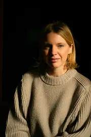 Portrait of a woman with blonde hair wearing a beige sweater against a dark background.