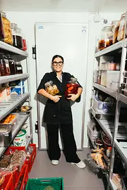 Chef standing in a storage room between shelves holding two large jars of pickled vegetables.