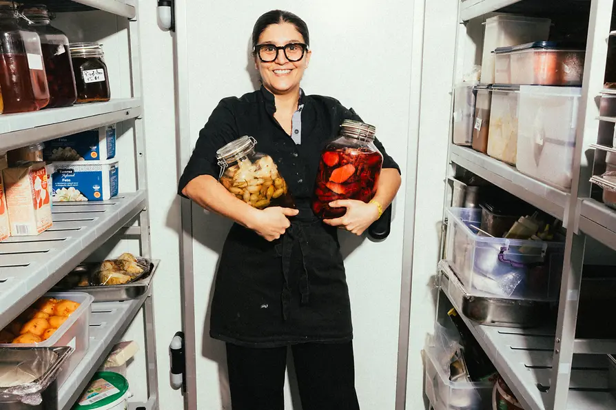 Köchin steht in einem Vorratsraum zwischen Regalen und hält zwei große Gläser mit eingelegtem Gemüse.
