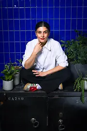 Woman sitting in front of a blue tiled wall surrounded by plants, looking at the camera.
