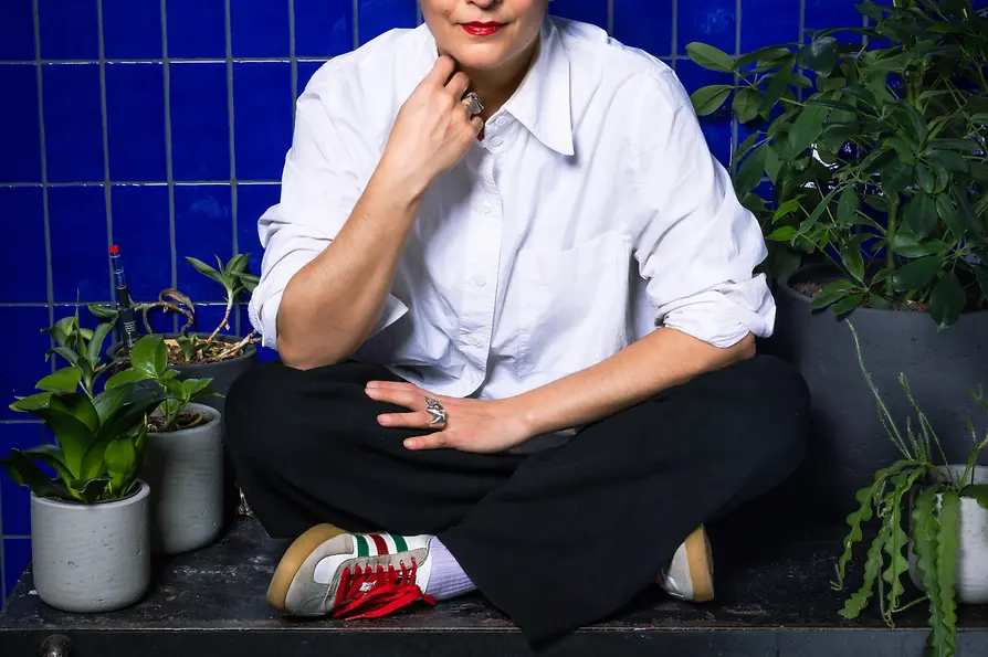 Woman sitting in front of a blue tiled wall surrounded by plants, looking at the camera.