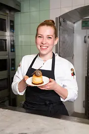 Pastry chef smiling in a bakery kitchen while presenting a small cake on a plate.