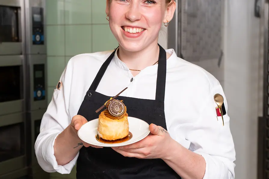 Pastry chef smiling in a bakery kitchen while presenting a small cake on a plate.
