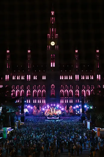 Crowd gathered before a stage on City Hall Square, the building illuminated in pink.