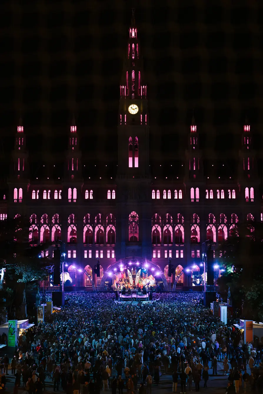 Crowd gathered before a stage on City Hall Square, the building illuminated in pink.