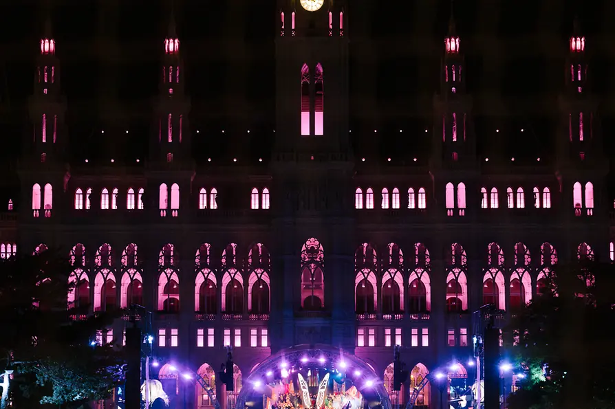 Crowd gathered before a stage on City Hall Square, the building illuminated in pink.