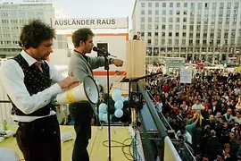 Speakers stand on a container addressing a large crowd beneath a political banner.