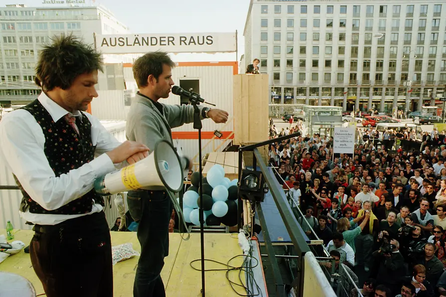 Speakers stand on a container addressing a large crowd beneath a political banner.