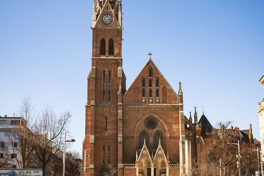 Neo-Gothic brick church with tall spire and clock against a clear blue sky.