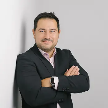 Mario Galler, General Manager of the Hotel Zeitgeist Vienna, business portrait. Standing, slightly leaning against a white wall, arms crossed, smiling warmly.