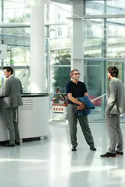 A congress attendee is standing at an information counter to register. Two other attendees are chatting next to them in a relaxed posture. All are wearing business attire. VIECON – Vienna Congress & Convention Center.