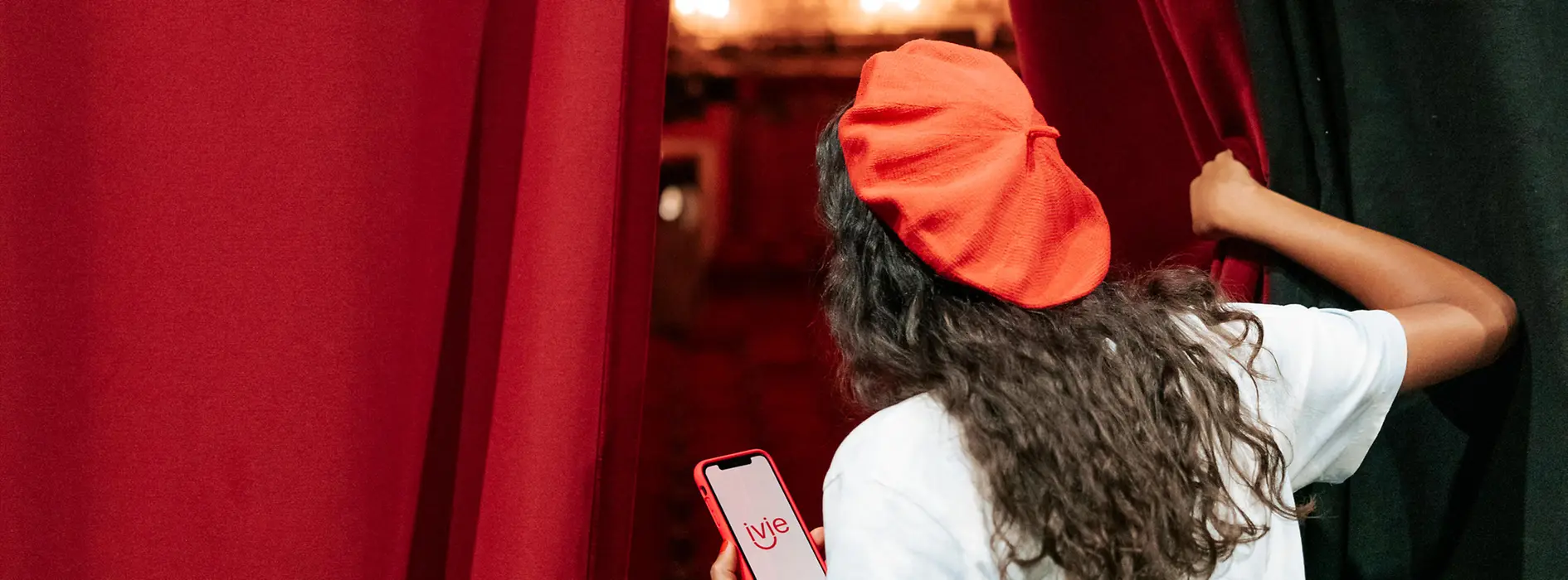 A woman stands behind a red curtain on the stage of the Vienna State Opera. With her right hand, she pulls the curtain aside and looks out toward the auditorium. In her left hand, she holds a mobile phone displaying the word “ivie.”
