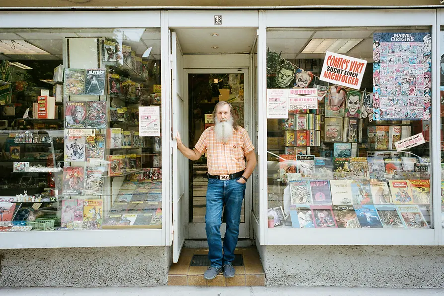 A man with a long white beard stands in the open doorway of a comic shop, with display windows packed with comics and posters.