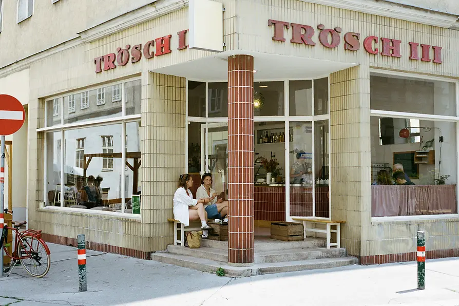 Corner café labeled “Trösch II” with large windows and two seated guests outside the entrance on a quiet street.