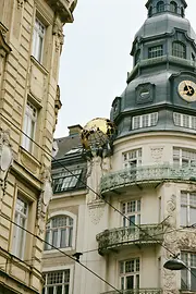 Verzierte Wiener Gründerzeitfassade mit Eckturm, Uhr und Figuren, die eine goldene Weltkugel über einem Balkon tragen.