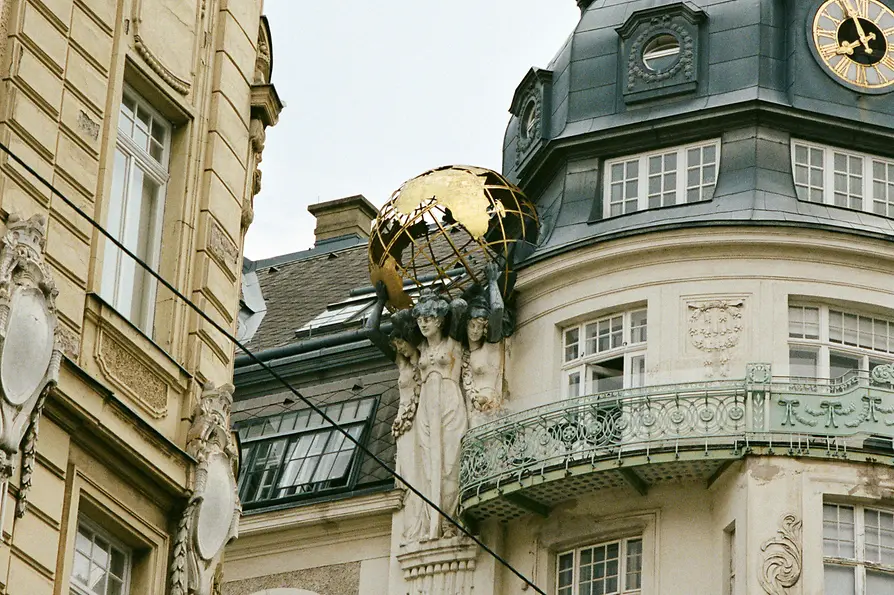 Verzierte Wiener Gründerzeitfassade mit Eckturm, Uhr und Figuren, die eine goldene Weltkugel über einem Balkon tragen.