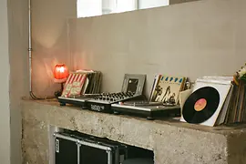 Plattenspieler und Mischpult stehen auf einem Betonregal neben gestapelten Schallplatten; eine kleine rote Lampe leuchtet am Fenster.