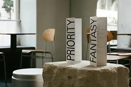 Zwei aufgestellte Menükarten mit den Worten „PRIORITY“ und „FANTASY“ stehen auf einem groben Steinblock im Caféraum.