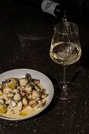 Weißwein wird in ein Glas eingeschenkt; daneben steht ein Teller mit Venusmuscheln in cremiger Sauce auf dunklem Tisch.