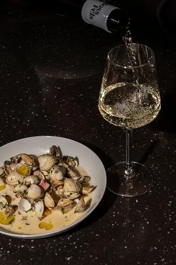 White wine is poured into a glass next to a plate of clams in a creamy sauce on a dark table.