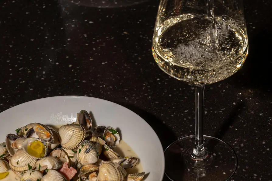Weißwein wird in ein Glas eingeschenkt; daneben steht ein Teller mit Venusmuscheln in cremiger Sauce auf dunklem Tisch.