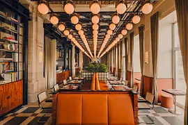 Langer Restaurant-Speisesaal mit orangenen Sitzbänken, gedeckten Tischen und vielen runden Hängeleuchten entlang der Decke.