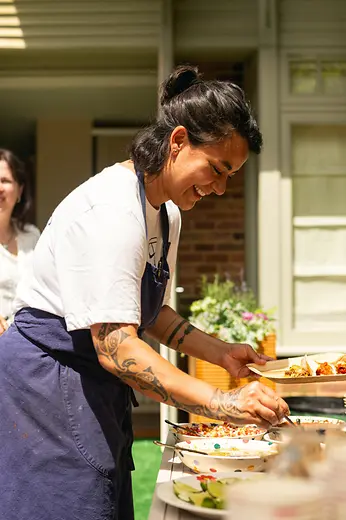 A chef in an apron arranges food at an outdoor table, plating a dish.