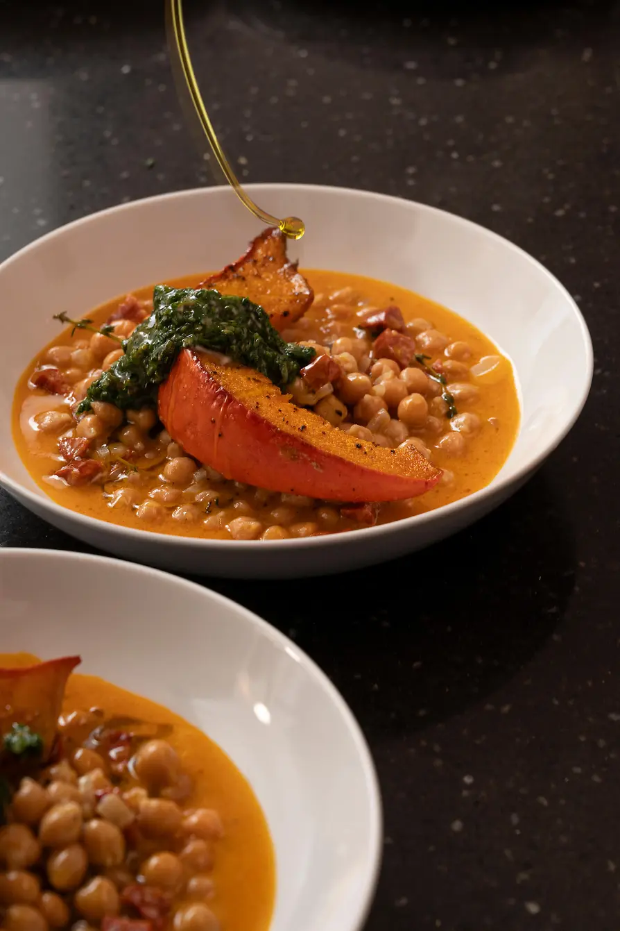 Chickpeas in a creamy sauce with roasted squash and herbs, with oil being drizzled into the bowl.