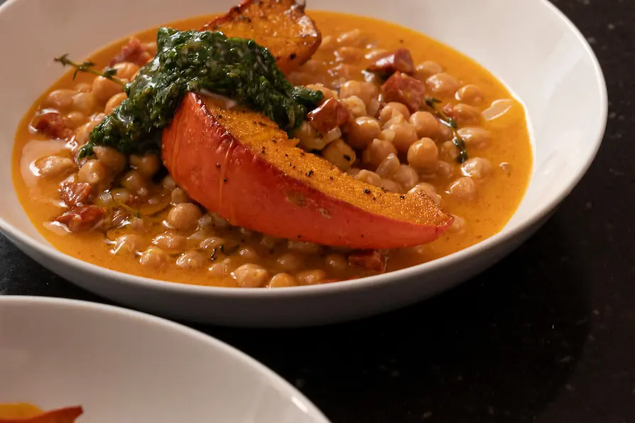 Kichererbsen in cremiger Sauce mit geröstetem Kürbisspalten und Kräutern; darüber wird Öl in die Schale geträufelt.