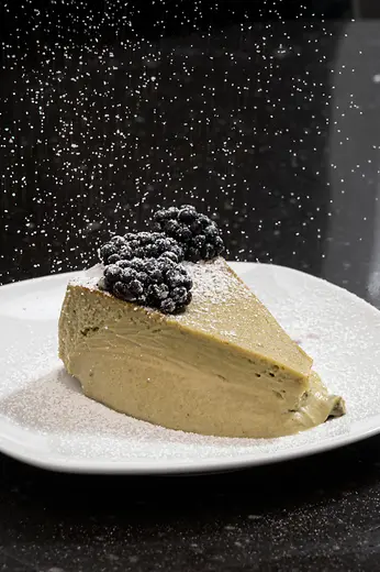 A slice of cheesecake topped with berries on a white plate as powdered sugar falls from above.