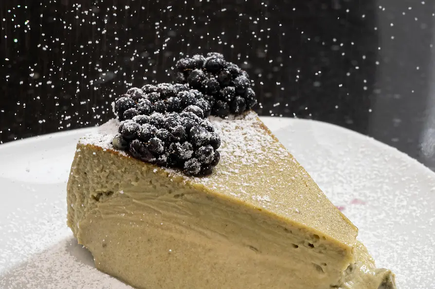 Stück Cheesecake mit Beeren auf einem weißen Teller, während Puderzucker von oben herabgestäubt wird.