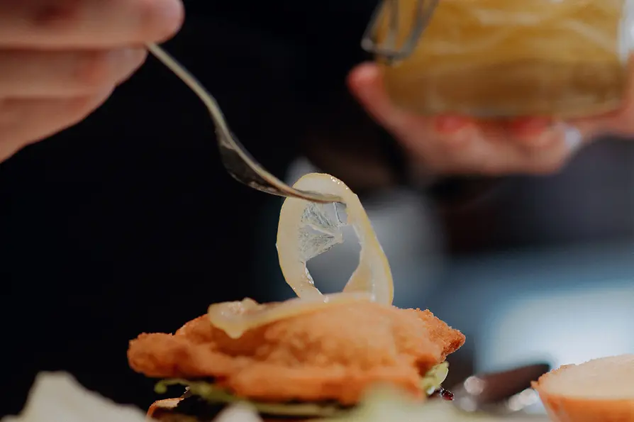 Mit einer Gabel wird ein Stück eingelegte Zitrone auf ein paniertes Schnitzel im Burger gelegt; im Hintergrund ein Glas mit Zitronen.