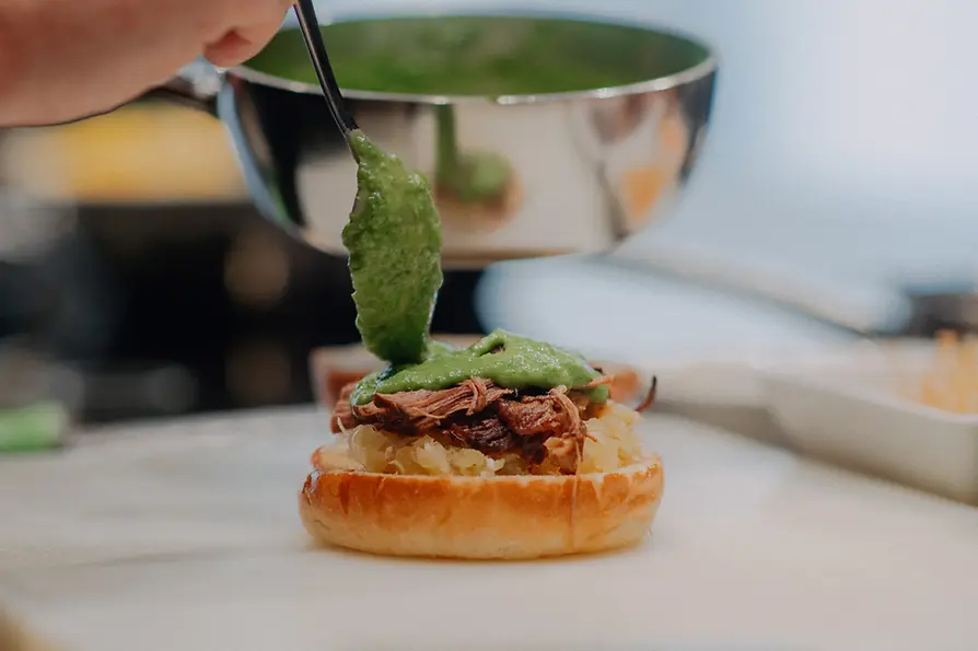 Eine Hand löffelt grüne Schnittlauchsauce aus einer Metallschüssel auf einen Burger mit Fleisch und Kraut.