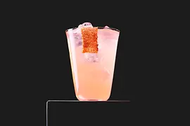 Rosafarbener Longdrink mit Eis steht auf einem Podest vor schwarzem Hintergrund; am Glasrand hängt ein Streifen Gewürzrand.