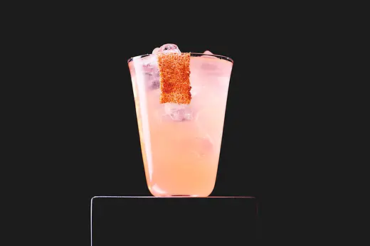 A pink iced long drink sits on a plinth against a black background, with a strip of spiced rim on the glass.