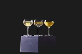 Drei Cocktails in Coupette-Gläsern stehen auf einem dunklen Podest vor schwarzem Hintergrund; in den Drinks liegen unterschiedliche Garnituren.