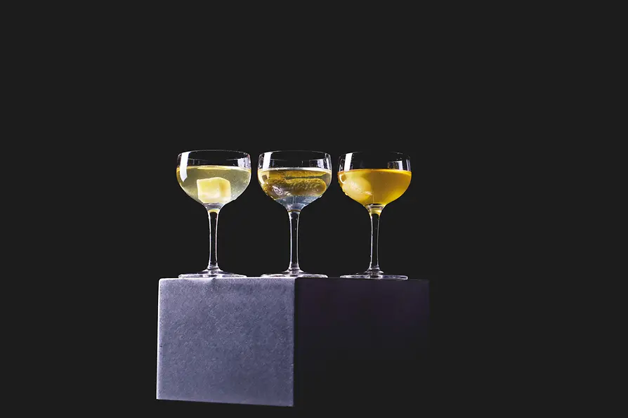 Drei Cocktails in Coupette-Gläsern stehen auf einem dunklen Podest vor schwarzem Hintergrund; in den Drinks liegen unterschiedliche Garnituren.