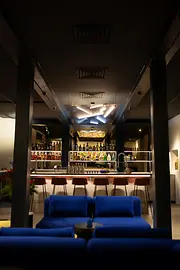 Blick aus dem Lounge-Bereich mit blauen Sofas auf eine helle Bar mit Flaschenregalen und leuchtender Deckeninstallation.