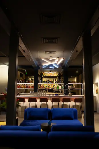View from a lounge area with blue sofas toward a bright bar lined with bottle shelves and a glowing ceiling installation.