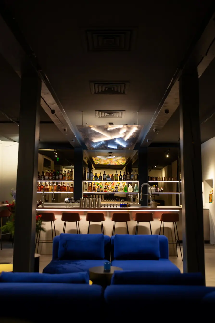 View from a lounge area with blue sofas toward a bright bar lined with bottle shelves and a glowing ceiling installation.