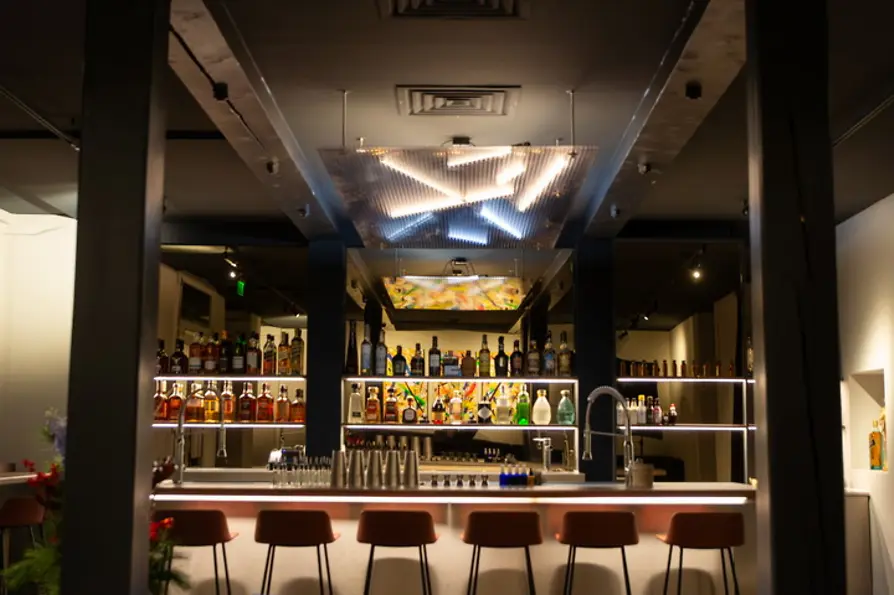 Blick aus dem Lounge-Bereich mit blauen Sofas auf eine helle Bar mit Flaschenregalen und leuchtender Deckeninstallation.