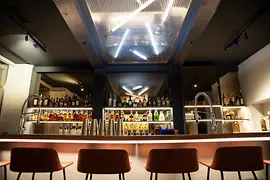 Moderne Bar mit beleuchteten Spirituosenregalen, Barhockern und glänzender Theke; darüber hängen geometrische Neonleuchten.