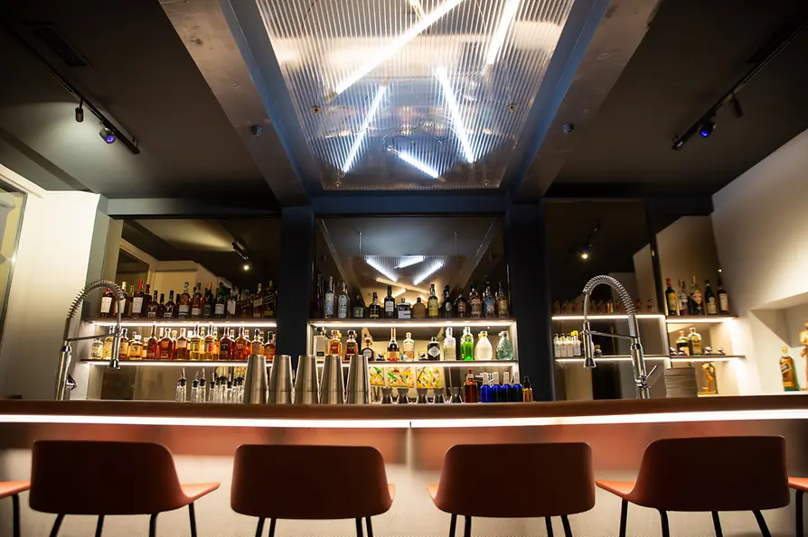Moderne Bar mit beleuchteten Spirituosenregalen, Barhockern und glänzender Theke; darüber hängen geometrische Neonleuchten.
