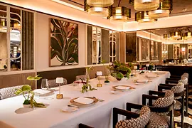 Lange festlich gedeckte Tafel mit Blumenarrangements und kleinen Lampen in einem eleganten Speisesaal mit Spiegeln und Kronleuchtern.