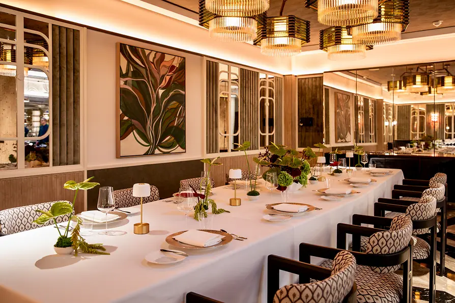 Lange festlich gedeckte Tafel mit Blumenarrangements und kleinen Lampen in einem eleganten Speisesaal mit Spiegeln und Kronleuchtern.